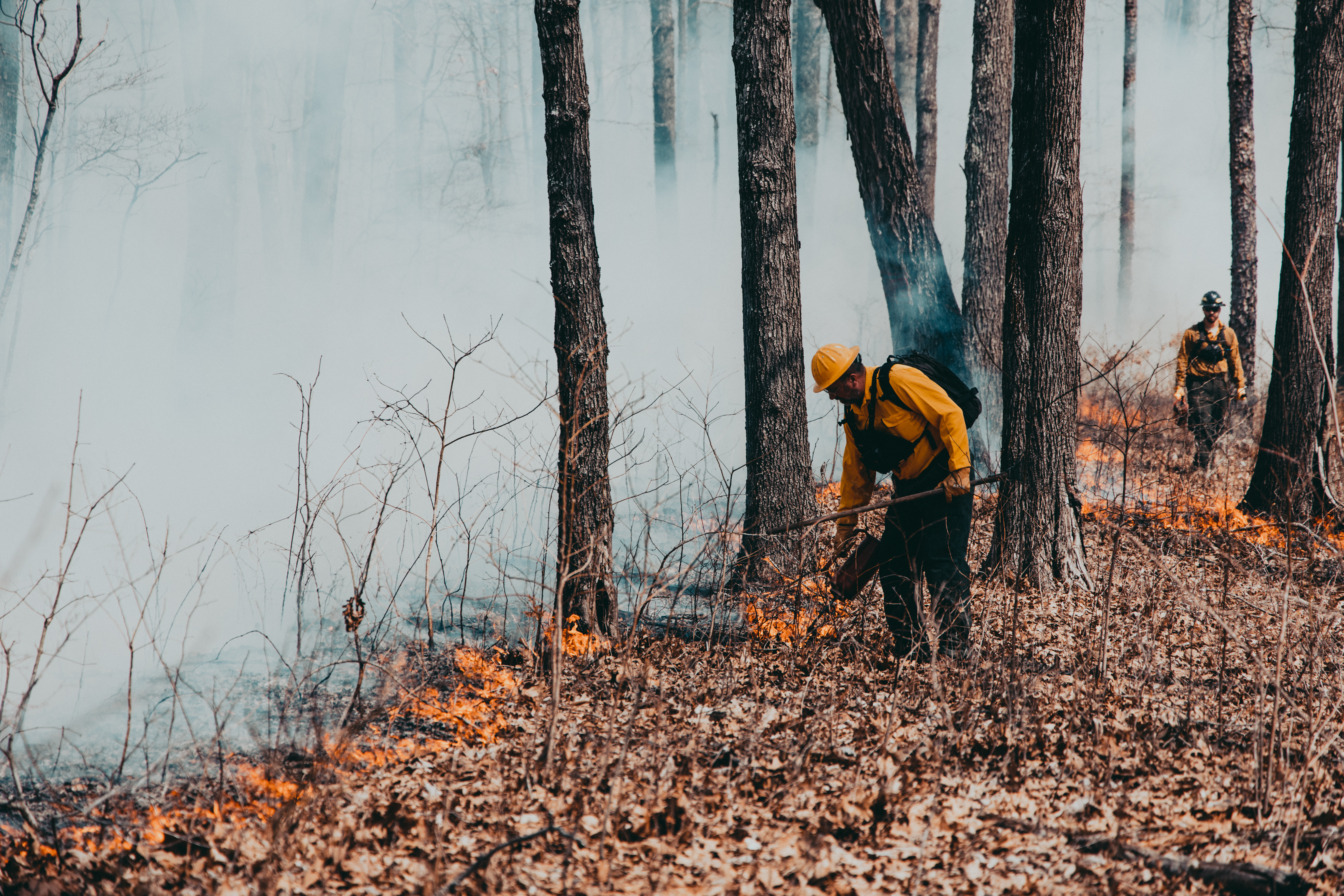 "Two firefighters walk along a wall of smoke, setting a low fire behind them.